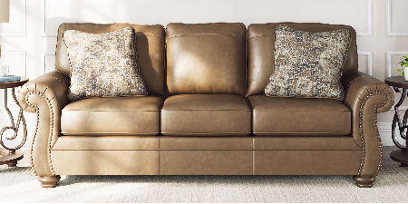 A brown leather loveseat sofa with rolled arms and nailhead trim, styled with two patterned throw pillows in a bright living room. Above the sofa hangs a rectangular canvas painting of birch trees in an autumn forest.